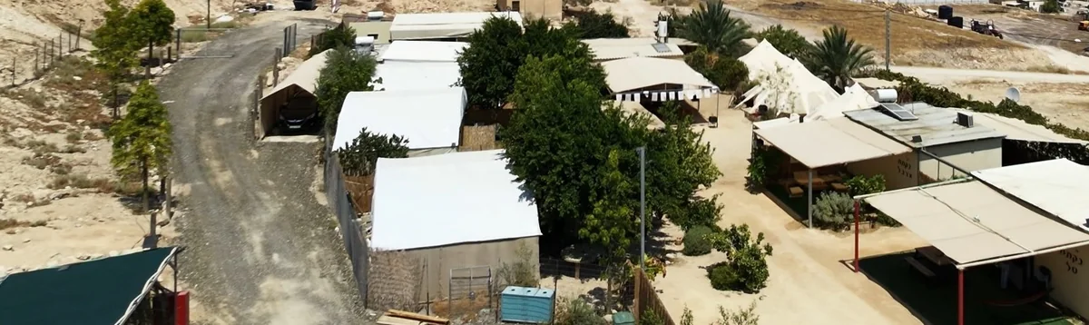מדברתא מהאוויר — דרך עפר יורדת במדרון המדברי אל האוהלים הלבנים והעצים